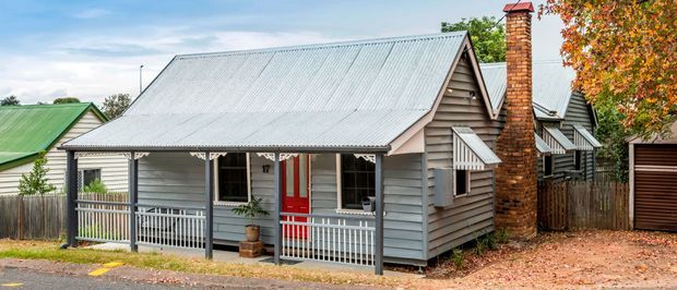 Street-facing view of a compact single-storey cottage with grey weatherboard cladding, a corrugated metal gable roof, white trim and porch railings, a bright red front door, and a prominent brick chimney. Autumn foliage and neighbouring homes frame the residential setting.