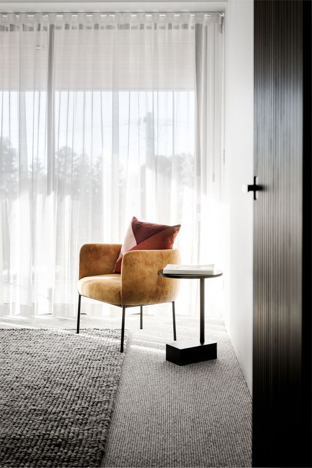 A bright, refined interior vignette featuring a caramel-brown upholstered armchair with a rust-toned cushion beside a small black pedestal side table. Sheer white curtains provide abundant filtered daylight, while a textured grey area rug and vertically ribbed dark wall panel add tactile contrast.