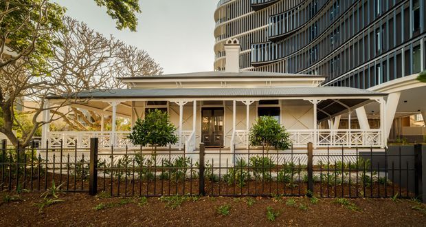 Street-facing view of a restored traditional timber house with a broad covered veranda, decorative white fretwork, pale weatherboard walls, and a low pitched roof. Mature trees and a landscaped front garden sit behind a metal fence, while a tall contemporary apartment building rises immediately behind the house, highlighting an urban heritage context.