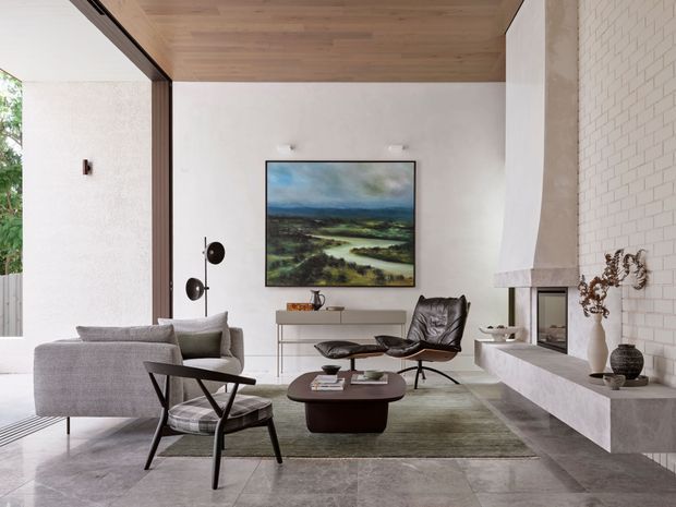 Refined contemporary living room with a grey sofa, black leather lounge chair, sculptural coffee table, and large landscape artwork. A full-height glazed opening brings in natural light beside a warm timber-lined ceiling, while a white brick fireplace wall with a substantial stone hearth forms the main architectural feature. The room has a spacious, calm, neutral-toned composition with a mix of timber, stone, plaster, and metal finishes.