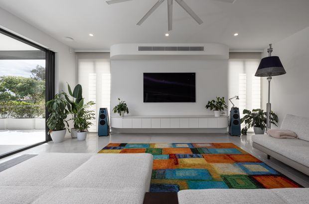 Bright contemporary living room with a white built-in media wall, wall-mounted television, integrated cabinetry, large sectional seating, colourful geometric area rug, indoor plants, floor-to-ceiling glazing, and a ceiling fan. The space has a clean, airy arrangement with strong colourful accents.