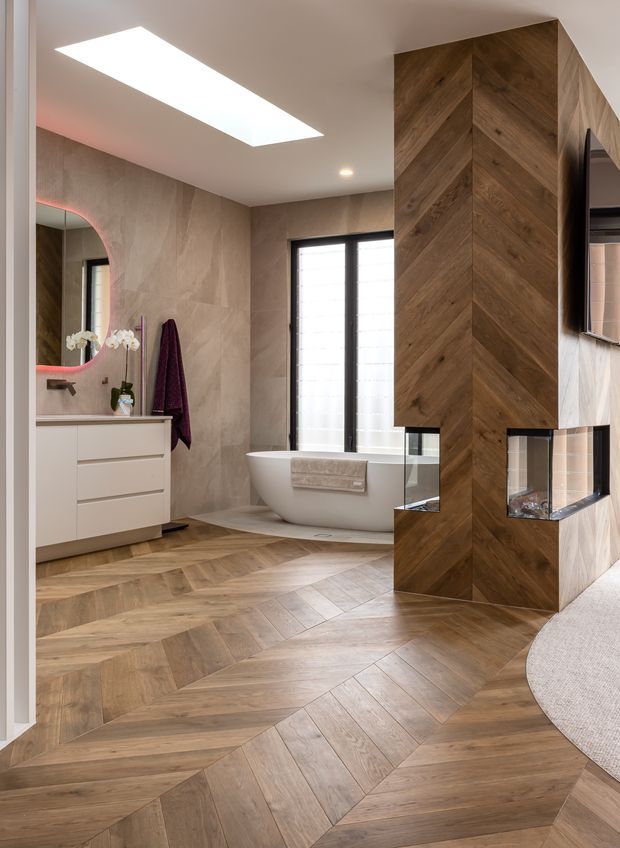 Contemporary bathroom with a freestanding white bathtub, floating white vanity, large-format stone-look wall tiles, black-framed window, illuminated rounded mirror, and warm timber chevron flooring. A substantial chevron timber-clad partition incorporates a recessed fireplace or display feature and partially screens the adjacent room.