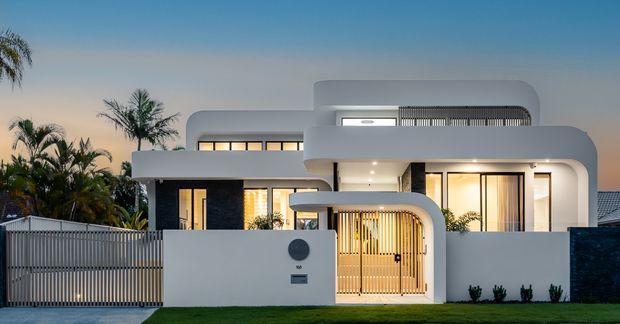 Contemporary two-storey residence photographed at dusk, featuring smooth white rendered walls, rounded corners, layered flat roof forms, expansive dark-framed glazing, and a sheltered entry framed by a curved portal and vertical timber-look screening. A minimalist front fence, landscaped planting, and warm interior lighting reinforce the refined modern facade.
