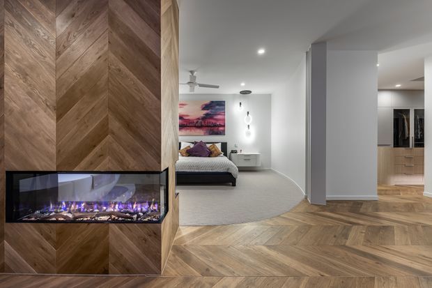 Contemporary open-plan interior featuring a dramatic herringbone timber feature wall with an inset linear electric fireplace. The timber herringbone flooring continues through the hallway and living area, leading toward a bedroom with a curved rug, upholstered bed, and colourful artwork. Recessed ceiling lighting, a ceiling fan, white walls, and integrated storage create a clean, refined residential interior.