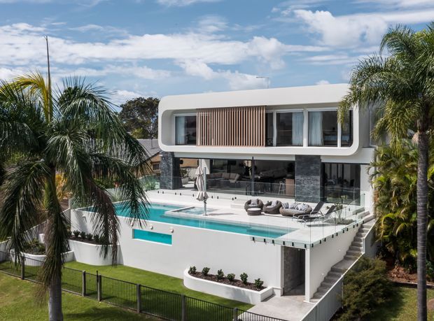 A contemporary multi-level residence viewed from the exterior, featuring a rounded white upper-storey volume with extensive glazing and vertical timber screening. Dark stone-clad walls frame an open living area overlooking a turquoise swimming pool and terrace, with stairs descending through landscaped grounds and palm trees surrounding the property.