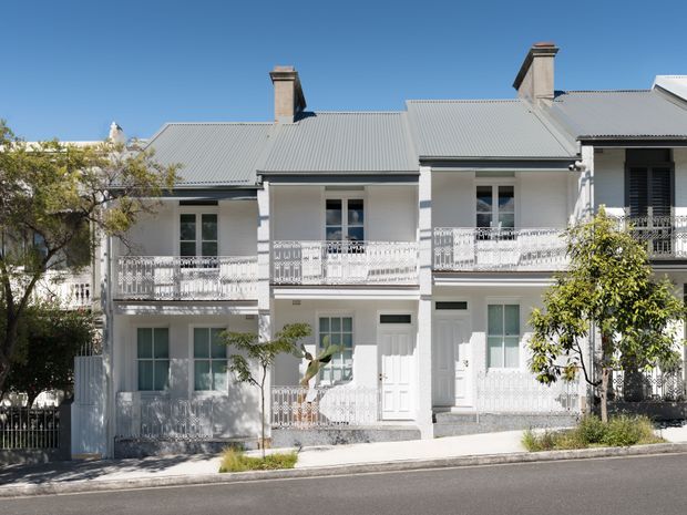 Street-facing view of a bright, multi-unit heritage-style residential building with a symmetrical white facade, ornate metal verandah and balcony balustrades, tall sash-like windows, multiple entry doors, and a broad corrugated metal roof with chimneys. Mature street trees partially frame the facade.