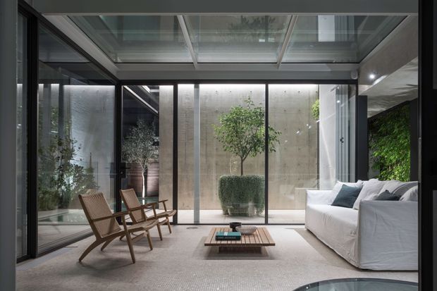 A refined, light-filled glass-enclosed living room arranged around a low timber coffee table. Full-height glazing and a glazed roof look onto a planted courtyard with concrete walls and greenery. The room includes a white slipcovered sofa, woven timber lounge chairs, a textured neutral rug, and dark-framed glass doors.