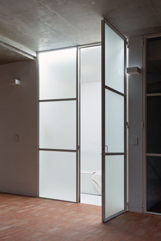 A minimalist interior showing a partially open frosted-glass door and adjoining frosted-glass partition panels in dark metal frames. The space has a raw concrete ceiling, plain light-coloured walls, and a red terracotta brick-tile floor. A glimpse of a tiled bathroom with a white towel is visible beyond the door.