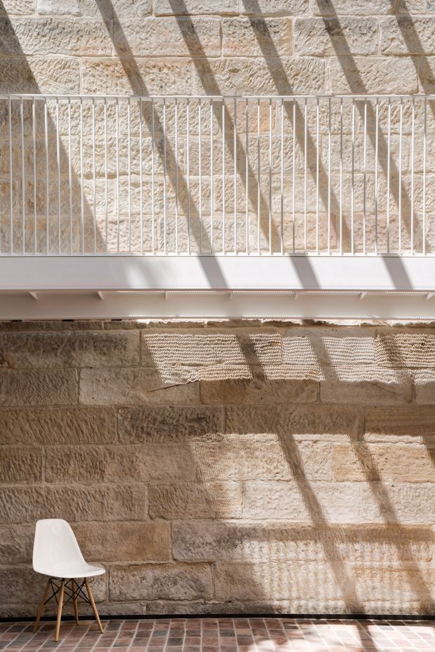 Sunlit interior with exposed warm sandstone block walls, a white metal mezzanine or balcony railing, and a high clerestory-like opening casting strong linear shadows. The space has a reddish brick or terracotta tiled floor and a single white molded chair with timber legs. The composition emphasises textured masonry, daylight, and dramatic geometric shadows.