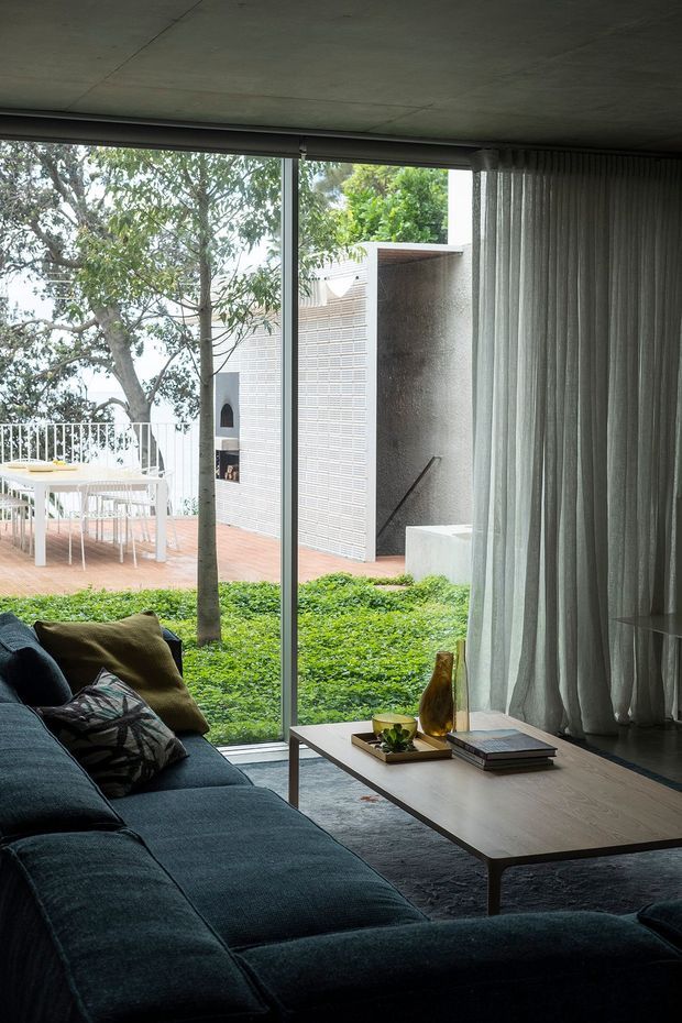 A contemporary living room with a large dark sectional sofa, low rectangular timber coffee table, patterned cushions, floor rug and full-height glazed doors or windows opening visually onto a planted courtyard terrace. Sheer curtains soften the glazing. Outside, a white brick garden wall, built-in outdoor fireplace and steps are visible, with a dining setting on a terracotta-paved terrace. The interior has a dark, intimate palette contrasted by abundant greenery and daylight.