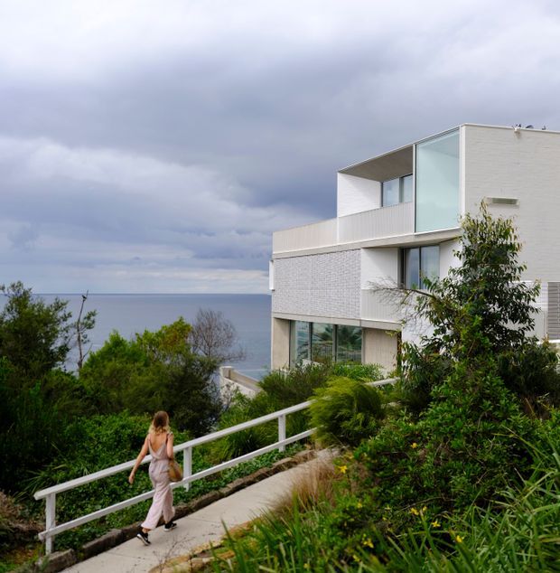 Exterior view of a modern, multi-storey coastal residence with white masonry walls, extensive glazing, and projecting flat-roofed forms. The house is set among dense landscaped vegetation on a sloping site, with a paved pedestrian path and ocean visible beyond.