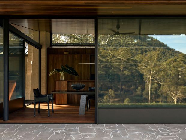 A refined, dark-toned interior viewed through expansive floor-to-ceiling glazing, featuring a timber-lined ceiling and floor, a rich dark timber built-in cabinet and bar joinery, a sculptural console table, and a black leather chair. The glazing reflects and frames a wooded landscape, while irregular grey stone paving forms the foreground. The room has a strong connection to nature and a sophisticated, minimalist character.