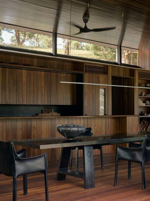 A warmly lit open-plan kitchen and dining area with extensive dark timber panelling and cabinetry. A long island with an integrated dark benchtop separates the kitchen from a dining table with sculptural timber legs and black leather armchairs. High clerestory-style windows bring in views of trees and surrounding landscape, while a dark ceiling fan and linear pendant light add functional detail. The space has a refined, nature-connected mid-century character.