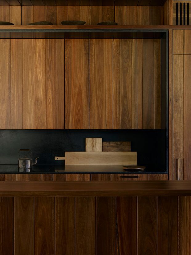 A tightly framed, dark-toned kitchen or bar joinery composition dominated by vertically panelled natural timber cabinetry and a timber-fronted counter. A black backsplash and dark shelving create strong contrast, with a small metal kettle, cutting boards, and understated decorative objects arranged on the work surface. The design is highly integrated and minimalist, with warm walnut-like timber and concealed or flush cabinetry.