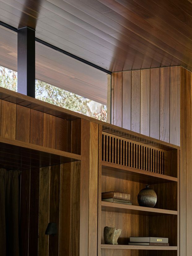 Close-up interior view of a richly timber-lined room featuring floor-to-ceiling built-in shelving and cabinetry, a slatted ventilation or decorative panel, and a dark structural post. Warm wood wall and ceiling panelling create a refined, intimate atmosphere, with a high clerestory-like glazed opening bringing in natural light. Shelves display books and decorative objects.