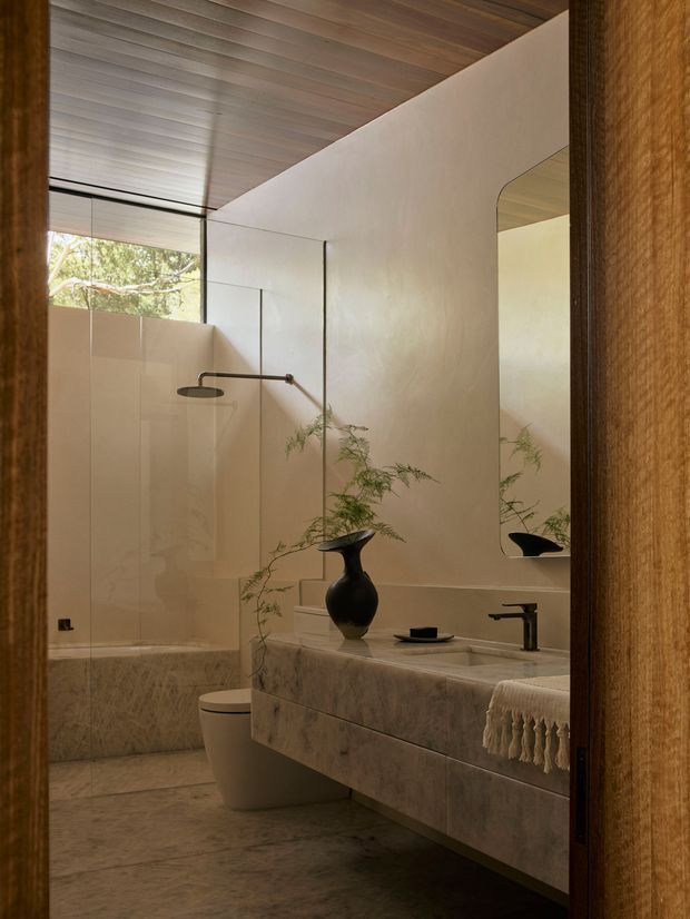 A warm, refined bathroom viewed through a timber-framed doorway. The room features a floating stone vanity with integrated basin, dark tapware, a large rounded-corner mirror, a wall-mounted shower with frameless glass enclosure, a concealed toilet, and a timber-lined sloped ceiling. Natural light enters through high glazing, creating a calm earthy atmosphere.