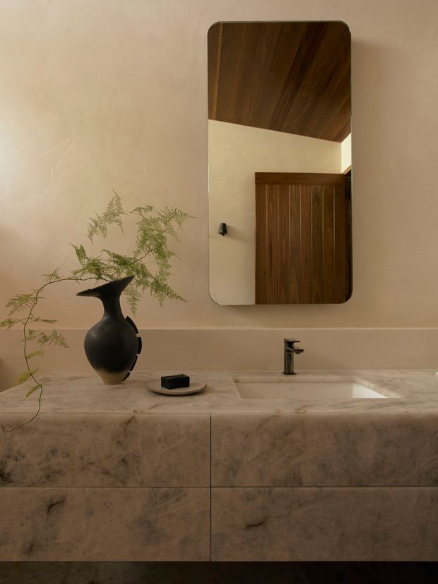 A warm, refined bathroom vanity scene with a full-width pale veined stone counter and integrated rectangular basin. A dark metal tap, sculptural black vase with delicate greenery, and soap dish sit on the counter. Above, a rounded-rectangle mirror reflects dark timber cabinetry and a sloped timber ceiling or wall, adding depth and contrast to the softly textured beige plaster wall.