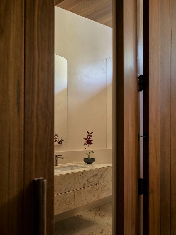 A refined bathroom viewed through a partially open timber door. The space features a floating stone vanity with integrated drawers, a pale marble or stone benchtop and matching wall panels, a wall-mounted basin tap, an organic-shaped mirror, and warm timber ceiling lining. Soft natural light and a small potted orchid create a calm, understated atmosphere.