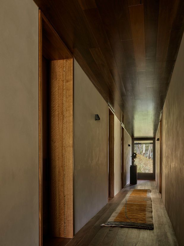 A long, narrow interior hallway with warm dark timber lining across the ceiling and door frames, smooth earthy plaster walls, and timber flooring. A textured runner leads toward a glazed door at the end, with a dark sculptural object silhouetted nearby. The space is dim, warm, and strongly material-focused, with minimal furnishings and concealed or subtle lighting.