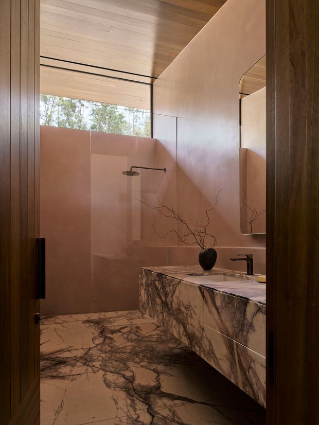A refined contemporary bathroom viewed through a dark timber doorway. The room features a floating marble vanity with an integrated basin and dark tapware, matching dramatic veined marble flooring, warm blush-toned wall panels, a frameless shower area, a large rounded-corner mirror, and a timber-lined ceiling. Natural light enters through high glazing, creating a warm, luxurious atmosphere.