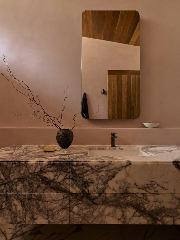 A warm, moody bathroom vanity with a full-width dramatic veined stone countertop and matching slab-front cabinetry. The room features a rectangular rounded-corner mirror, black tapware, a white integrated basin, muted terracotta plaster walls, and a dark timber ceiling reflected in the mirror. Minimal sculptural accessories and branch-like decor add an organic accent.