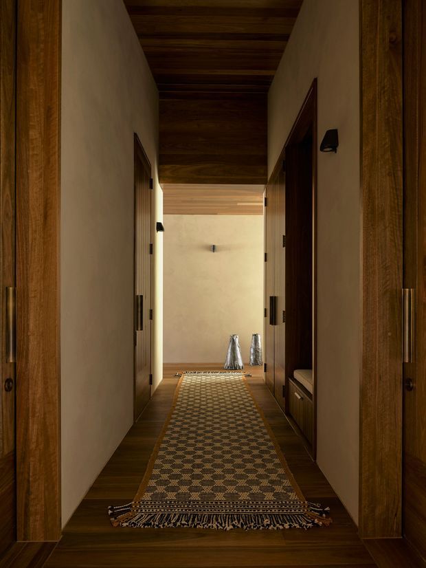A narrow, warm-toned residential hallway viewed through timber-framed openings. The corridor has dark timber flooring, a dark exposed timber plank ceiling, smooth cream plaster walls, timber doors and built-in joinery, and a long patterned runner rug leading toward a softly lit end wall. Small wall lights and decorative objects add subtle detail.