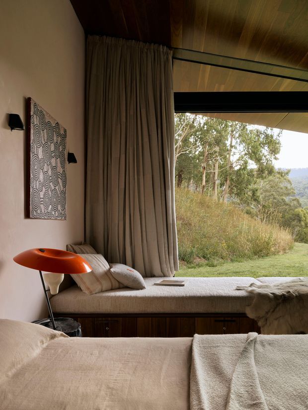 A warm, softly lit bedroom with a built-in window seat overlooking a wooded landscape. The room features a timber-lined ceiling, full-height taupe curtains, neutral upholstered bedding and cushions, dark timber storage cabinetry beneath the window seat, black wall sconces, and a distinctive orange mid-century-style floor lamp. Large glazing and an openable window create a strong connection to the natural setting.