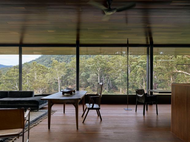 A refined living and dining space with expansive floor-to-ceiling glazing overlooking a wooded landscape. The room features warm timber flooring and ceiling boards, dark slender window frames, a low dark sofa, mid-century-inspired timber dining furniture, and a streamlined timber cabinet. A ceiling fan and soft blinds add practical detail, while the broad windows create a strong connection to the surrounding trees.