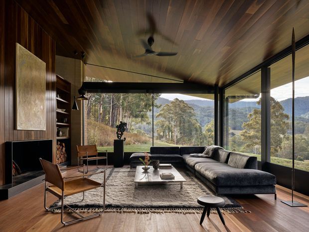A warm, wood-lined living room with a low dark sectional sofa, leather sling chairs, a central coffee table, built-in shelving and fireplace, and expansive floor-to-ceiling glazing overlooking a wooded mountain landscape. The room has a timber-clad ceiling and walls, dark metal window frames, layered pendant lighting, and a ceiling fan.