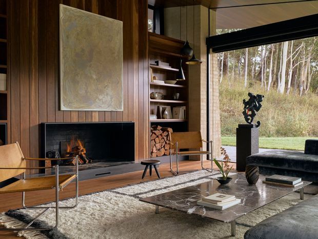 A sophisticated living room with a substantial open fireplace set into dark vertical timber wall panelling, integrated timber shelving and firewood storage, mid-century style lounge chairs, a dark sofa, and a low glass-topped coffee table. Large black-framed glazing opens the room to a wooded landscape. The space has a warm, refined, nature-connected atmosphere with strong mid-century architectural and furnishing cues.