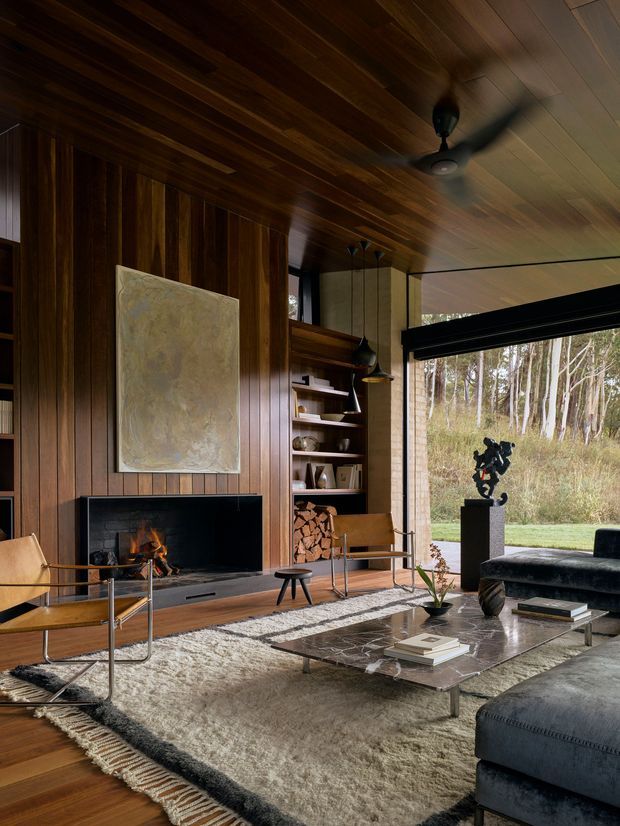 A richly detailed mid-century-inspired living room with a floor-to-ceiling dark timber feature wall and integrated open shelving surrounding a black fireplace. The room includes a low, glass-topped rectangular coffee table, textured area rugs, leather lounge chairs, a large dark upholstered sectional, sculptural decor, and a wood-panelled ceiling. Expansive glazing opens views to a wooded landscape, while a ceiling fan and pendant lights add functional and decorative detail. The atmosphere is warm, sophisticated, and inviting.