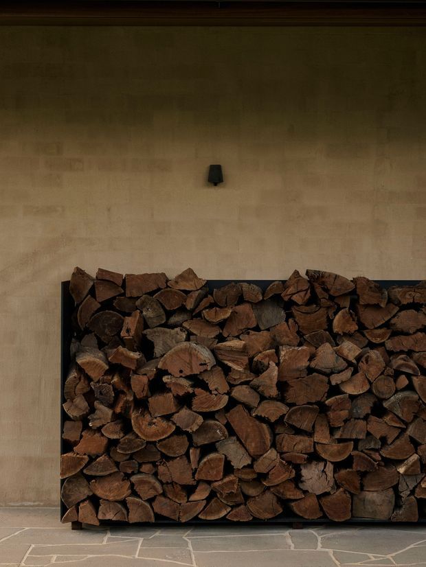 A moody close-up of a built-in fireplace wood-storage feature. Split firewood is neatly stacked within a dark rectangular metal frame against a warm earthy plaster wall, with a small black wall light above. The floor is finished in irregular pale stone slabs with contrasting grout lines. The image has a rustic, tactile and carefully composed architectural interior feel.