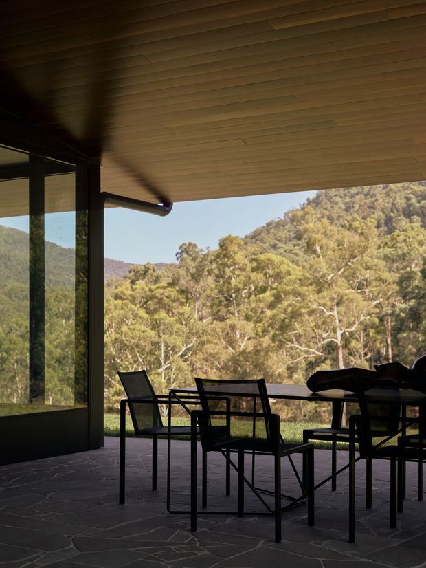 A covered outdoor dining terrace adjoining a glazed building, with a dark-framed corner window, timber-lined soffit, exposed guttering, and slate-like irregular stone paving. A dining table and chairs sit beneath the overhang, overlooking a wooded hillside landscape. The image has a strong indoor-outdoor connection and a shaded, warm, nature-oriented atmosphere.