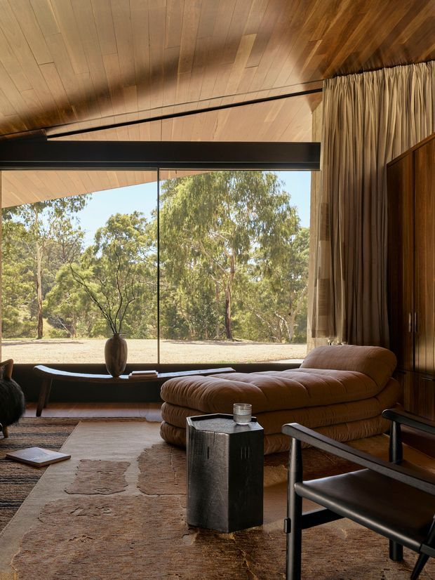 Warm, sophisticated living room with a low brown leather chaise lounge, black-framed floor-to-ceiling glazing, timber-lined sloping ceiling, dark timber built-in cabinetry, layered curtains, and a neutral patterned rug. The room opens visually to a wooded landscape and has a calm, refined mid-century-inspired aesthetic.