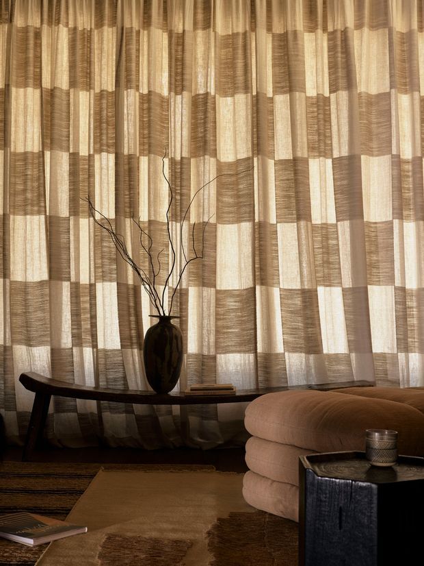 A warm, softly lit living room corner featuring full-height patterned sheer curtains in alternating beige and taupe panels, a dark sculptural console table with a black vase and branches, a low rounded beige upholstered sofa, and a dark side table. Layered neutral rugs and timber-toned flooring create a cozy, refined atmosphere.