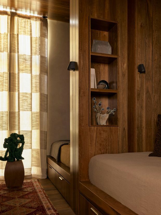 Warm, intimate bedroom interior with extensive dark timber wall panelling and built-in joinery. The room features a built-in bed with drawers, an integrated open display niche, a second bed visible beside the window, sheer woven curtains, a patterned rug, and warm low-level lighting. The design has a rich, cocooning, timber-lined character with compact built-in sleeping spaces.
