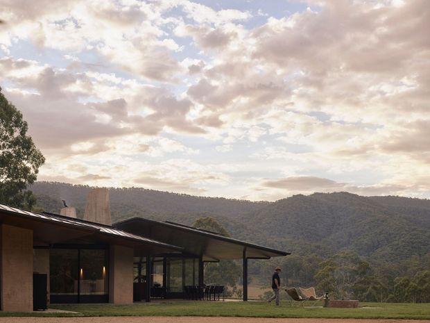 A low-slung contemporary home set against a densely forested mountainous landscape. The building has broad dark roof planes with deep overhangs, large areas of glazing, and warm earthy wall finishes. A covered outdoor terrace and lawn extend toward the view, with lounge furniture and a small outdoor fire feature visible near the edge. The image is photographed at dusk or in soft evening light beneath a cloud-filled sky.