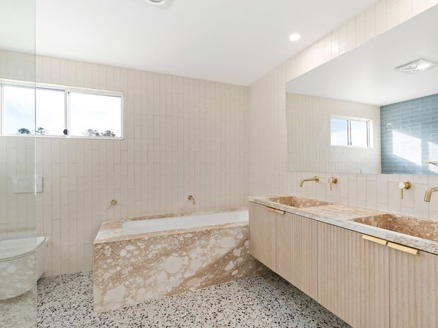 Bright, spacious bathroom with a freestanding-style built-in bath and double vanity. The room features pale vertical wall tiles, terrazzo flooring, a large mirrored wall, warm brass fixtures, and a blue-tiled shower area visible in the reflection. A toilet and shower enclosure are also visible at the left.