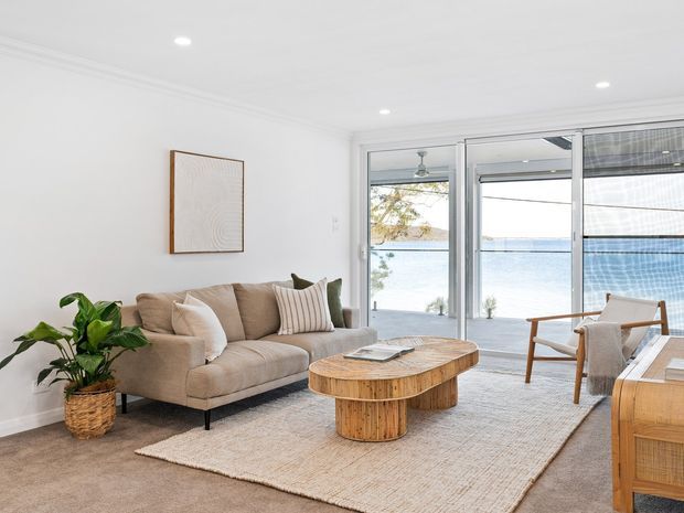 Bright, airy living room with a beige sectional sofa, textured neutral rug, rounded timber coffee table, woven planter, and timber-framed occasional furniture. Large sliding glass doors open to a waterfront view, bringing in abundant natural light. The room has white walls and ceiling, recessed lighting, simple cornice detailing, and a calm contemporary coastal feel.