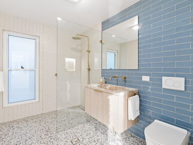 Bright contemporary bathroom with a walk-in glass shower, freestanding rainfall shower fittings, wall-mounted fluted vanity, large mirror, wall-hung toilet, and terrazzo-style flooring. The walls combine white herringbone tiles with blue rectangular tiles and brass-toned fixtures.
