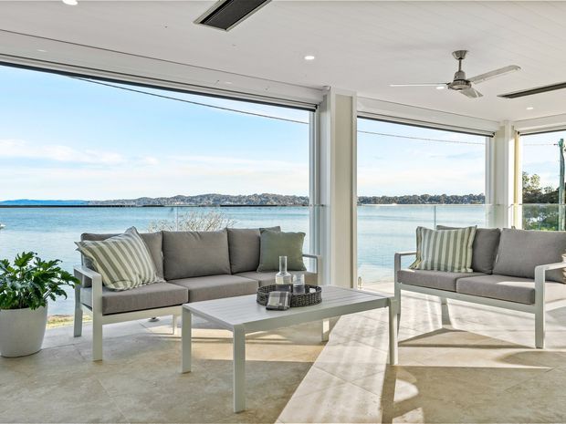 Bright waterfront living room with large floor-to-ceiling windows and wide glazed openings overlooking calm blue water and a distant shoreline. The room features two light grey upholstered sofas with striped cushions, a white rectangular coffee table, pale stone or tile flooring, white structural columns, and a white paneled ceiling with recessed lighting and a ceiling fan. The space has an airy, relaxed contemporary coastal character.