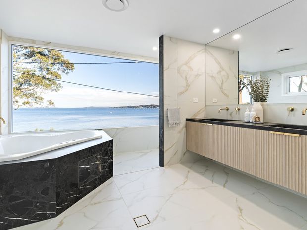 Luxurious contemporary bathroom with a freestanding white bathtub positioned beside expansive glazing overlooking the water. The room features large white marble-look floor and wall surfaces with grey veining, a black marble-look bath surround, and a floating fluted timber vanity with a dark benchtop and brass fittings. Recessed ceiling lighting and clean-lined detailing create a bright, refined atmosphere.