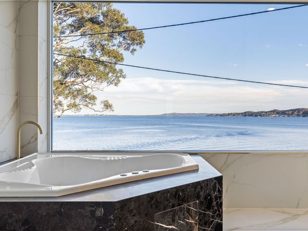 Luxury bathroom with a built-in spa bath positioned beside expansive glazing overlooking calm blue water and a distant shoreline. The room features white marble-effect wall surfaces, a dark veined stone bath surround, and a curved metallic tap. Reflections and power lines are visible across the waterfront view.