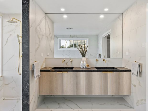 Bright contemporary bathroom with a double vanity, fluted timber-look cabinetry, black stone benchtop, large frameless mirror, and white marble-look wall and floor tiles. A walk-in shower with gold fixtures is visible at left, while a window and reflected bathroom elements appear in the mirror.