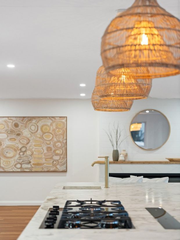 Bright, warmly styled kitchen with a large light stone island and inset gas cooktop in the foreground. Woven rattan pendant lights hang above the island, while a timber shelf, round wall mirror, abstract artwork, and neutral cabinetry or walls create a relaxed natural aesthetic.