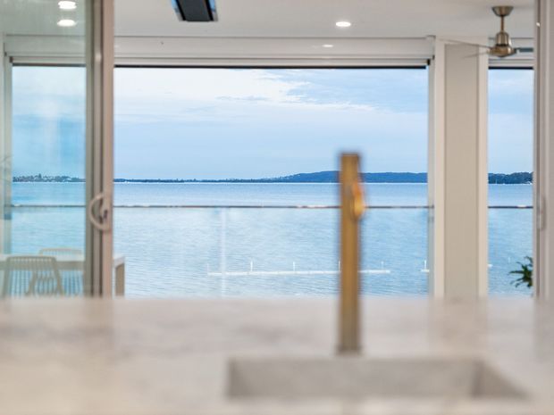 A bright, contemporary kitchen or open-plan living space viewed toward expansive waterfront glazing. The foreground shows a pale stone island benchtop with an undermount sink and tall metallic tap, while large sliding glass doors frame calm blue water and a distant shoreline. White structural columns, recessed ceiling lights, and a ceiling fan create a clean, airy interior.