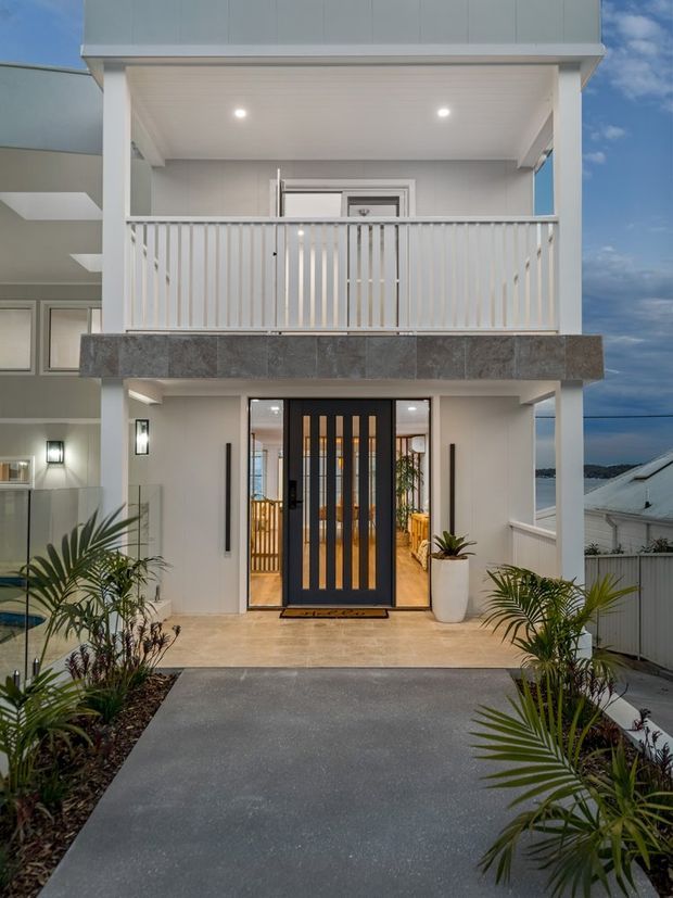 A dusk exterior view of a contemporary multi-level home, focused on the covered front entry and upper balcony. The facade combines white rendered walls, a dark framed glazed entry door, a white vertical-slat balcony balustrade, and a dark stone-clad balcony edge. A paved walkway and tropical planting lead to the entrance, with glimpses of neighbouring buildings and water beyond.
