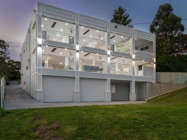 Twilight exterior view of a tall contemporary multi-storey residence on a sloping site. The building has a pale grey rendered facade, strong rectangular framing, extensive floor-to-ceiling glazing, illuminated interiors, projecting balconies, and an integrated lower-level garage or undercroft. Lawns, retaining elements, and neighbouring vegetation surround the property.