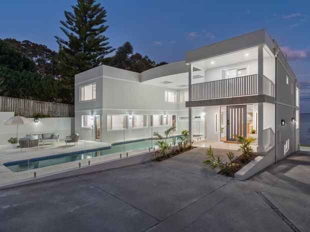 Twilight exterior view of a contemporary two-storey white residence with strong geometric massing, a flat roof, covered upper balcony with dark vertical balustrades, large glazed openings, and a recessed entry. A narrow swimming pool, paved outdoor area, landscaped planting, and adjacent driveway occupy the foreground, with trees and a glimpse of water beyond.