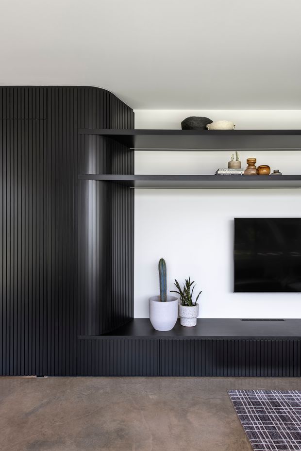 A minimalist living room media wall featuring extensive dark vertical fluted fixed joinery, open black display shelves, a wall-mounted television, and a low built-in console. White walls and concealed linear lighting provide contrast, while potted plants and small decorative objects add restrained styling. The room has a polished concrete floor and a dark patterned rug visible in the foreground.